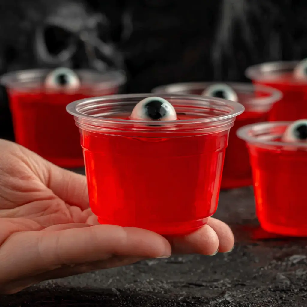 Halloween Eyeball Jello Shots: A Spooky Party Favorite 6 Hand holding a red jello cup with a candy eyeball on top, surrounded by more Halloween treats.